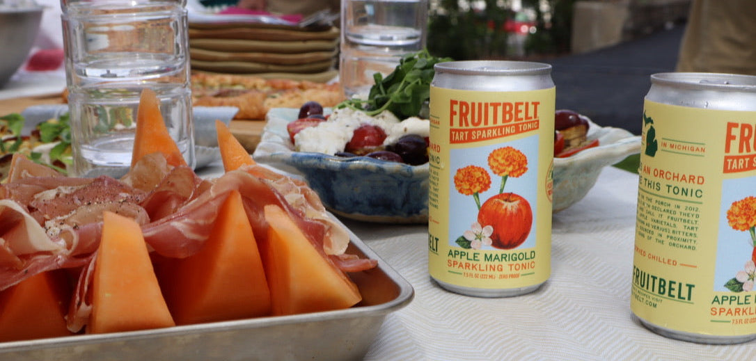 Two cans of Fruitbelt Sparkling Tonic apple marigold flavor are displayed on a white tablecloth next to a serving tray of prosciutto and cantaloupe melon slices, with outdoor dining elements in the background.