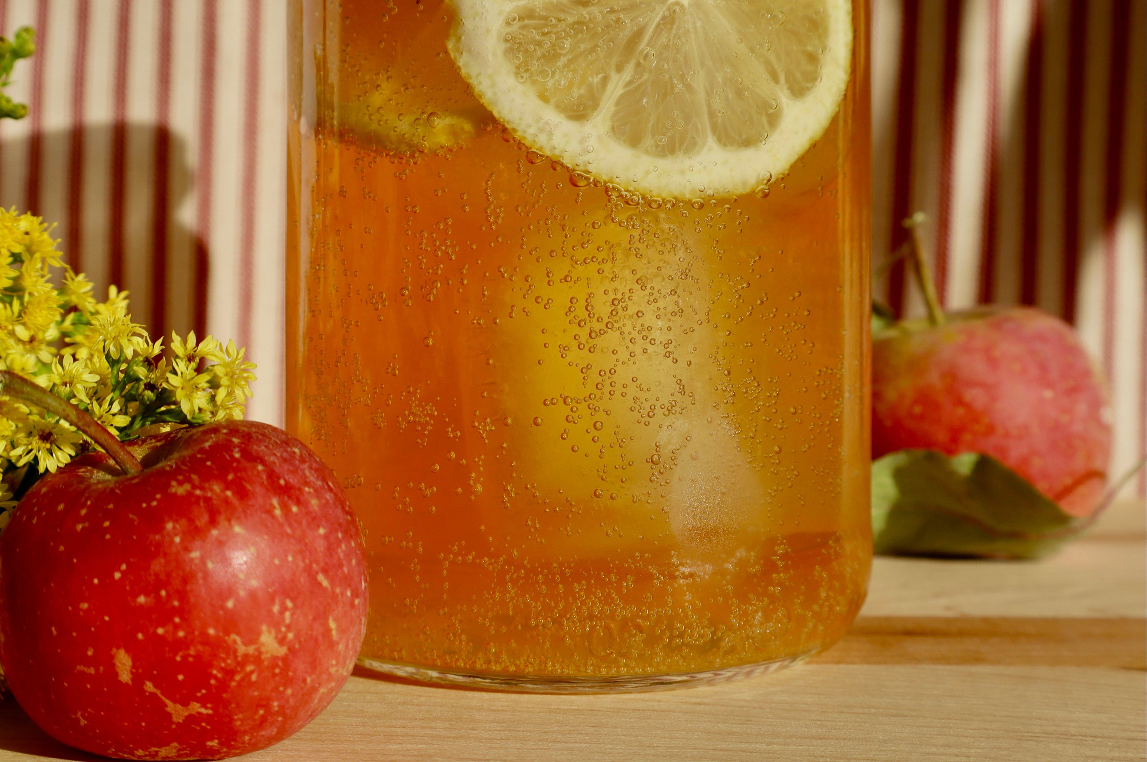 A clear glass pitcher filled with a golden iced beverage and carbon bubbles, garnished with a lemon slice, styled on a wooden surface with red apples and yellow flowers in warm sunlight.