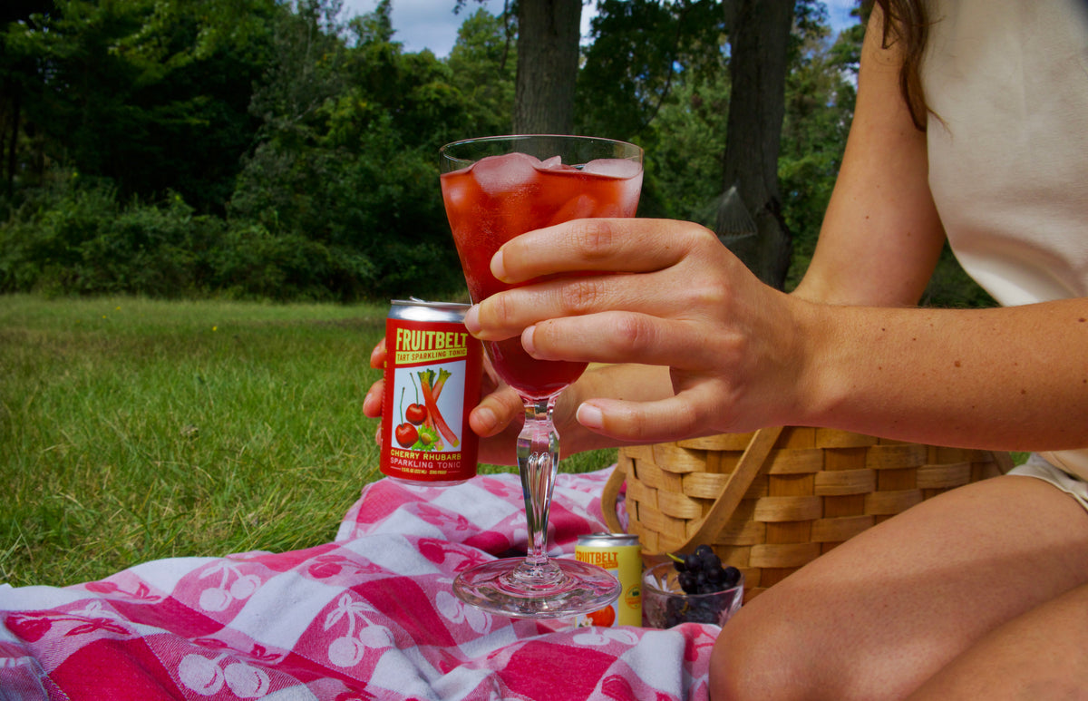 A person at an outdoor picnic holds a glass of red fruit punch next to a can of FruitBelt cherry rhubarb. The picnic setup includes a pink and white patterned blanket, a woven basket, and a green grassy field with trees in the background.