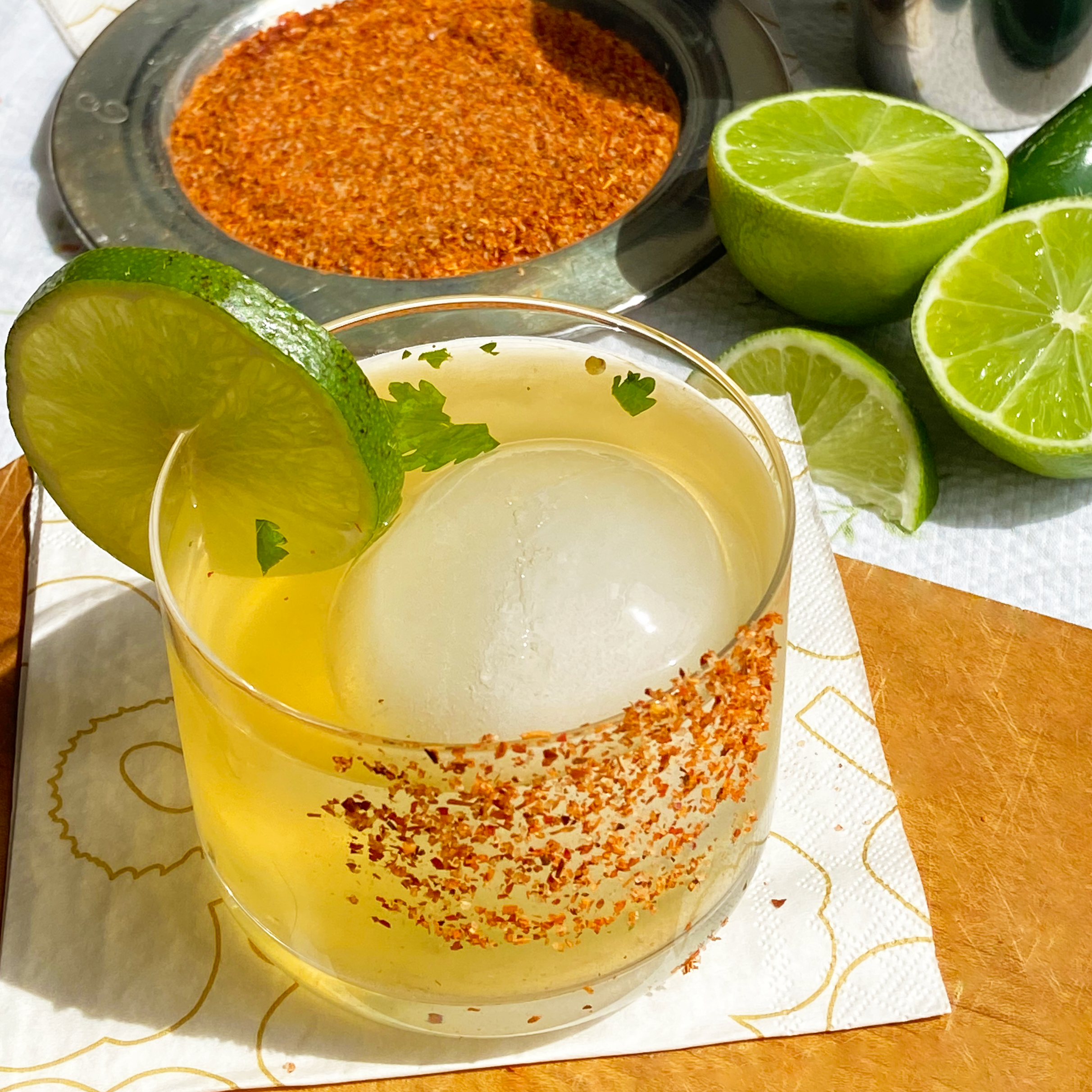 Glass of yellow liquid with ice and lime garnish on a wooden surface, surrounded by spices and limes.